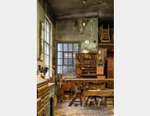 Alte Werkstatt mit hölzernen Werkbänken, Werkzeugregalen an der Wand und einer Sammlung von Handwerkzeugen. Der Raum hat abgenutzte Wände, eine Holzdecke und natürliche Beleuchtung durch ein großes Fenster.