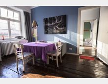 Wohnzimmer mit rustikalem Holzboden, einer blau gestrichenen Wand und einem Esstisch, der mit einer lila Tischdecke bedeckt ist. An der Wand hängt ein großes Naturfoto, und durch ein großes Fenster fällt Tageslicht in den Raum. Im Hintergrund ist ein angrenzender Raum mit einem weißen Sessel sichtbar.