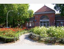 Backsteingebäude mit einem großen Bogenfenster in der Mitte und der Aufschrift „Sonnenallee“ über dem Eingang, umgeben von gepflegten Rosenbeeten und einer gepflasterten Wegführung; im Hintergrund stehen große Laubbäume.