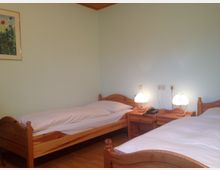 Ein einfaches Schlafzimmer mit zwei Einzelbetten aus Holz, dazwischen zwei Nachttische mit kleinen Tischlampen. Die Wände sind hellgrün gestrichen, und an der Wand hängt ein Bild mit Blumenmotiv.