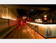 Innenraum einer modernen Bar mit Holzboden, einer langen beleuchteten Theke mit Barhockern und einer gepolsterten Sitzbank entlang der Wand. Die Wände zeigen kunstvolle Wandmalereien, und die Beleuchtung schafft eine warme, stimmungsvolle Atmosphäre.