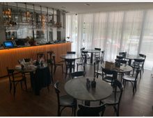 Ein modernes Restaurant mit dunklem Holzboden, runden Tischen mit gedecktem Geschirr und schwarzen Stühlen. Im Hintergrund befindet sich eine beleuchtete Bar mit einem Regal voller Gläser und Flaschen, während helle Vorhänge große Fenster bedecken.