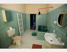 Ein einfaches Badezimmer mit türkisen Wandfliesen und weißen Bodenfliesen. Es enthält eine Toilette, eine dunkelgrüne Badewanne mit Duscharmatur, ein Waschbecken mit rundem Spiegel und einen Heizkörper in Handtuchhalter-Form.