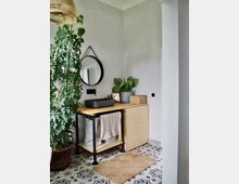 Minimalistisch eingerichtetes Badezimmer mit hellem Holzwaschtisch, schwarzem Aufsatzwaschbecken, rundem Wandspiegel und Pflanzendekoration. Der Boden hat ein schwarz-weißes Fliesenmuster, und ein Sisalteppich liegt davor.