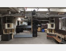 Ein Kellerraum mit improvisiertem Veranstaltungsbereich, ausgestattet mit großen Lautsprechern, einem DJ-Pult auf einem Teppich und diversen Kabeln und Geräten an den Wänden. Der Raum hat freiliegende Rohrleitungen an der Decke, unverputzte Wände und einfache Holzmöbel.