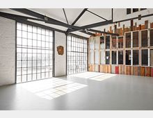 Ein leerer, industriell gestalteter Raum mit großen Stahlrahmenfenstern und weiß gestrichenen Ziegelwänden. An einer Wand sind alte Fensterrahmen und verschiedene Holzlatten angebracht, die als dekoratives Element dienen, während durch die Fenster viel Licht einfällt.