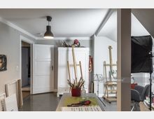 Ein Atelierraum mit neutral gestrichenen Wänden und Holzboden, ausgestattet mit Staffeleien, einem weißen Schrank, einem Tisch mit Malutensilien und einer Stehlampe. Im Hintergrund sind ein schwarzer Fotohintergrund und weitere Sitzgelegenheiten sichtbar.