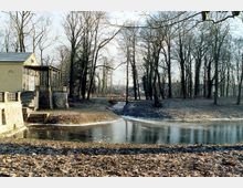 Parklandschaft mit einem zugefrorenen Teich im Vordergrund, kahlen Bäumen und einer kleinen Holzbrücke über einen Bach im Hintergrund; links ein historisches Gebäude mit einer steinernen Treppe und einem Balkon.