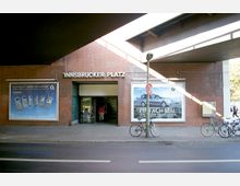 Eingang zur U-Bahn-Station "Innsbrucker Platz" in Berlin mit einer Klinkerfassade, flankiert von Werbeplakaten. Im Vordergrund stehen Fahrräder an einem Straßenpfosten, und oben verläuft eine erhöhte Brückenkonstruktion.