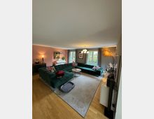 Wohnzimmer mit großer grüner Eckcouch, hellem Teppich und Holzboden. Die Wände sind teils rosa gestrichen, mit dekorativen Bildern, mehreren Fenstern und stilvollen Lampen, darunter ein Kronleuchter und eine goldene Stehlampe.