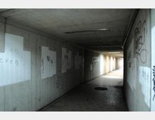 Betonierter Fußgängertunnel mit weißen Wänden, die teils Graffiti und übermalte Flächen aufweisen; der Tunnel ist von schwachem Licht beleuchtet und führt geradeaus in die Ferne.