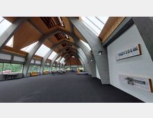 Großer Innenraum mit moderner Architektur, geprägt von einer gewölbten Holz- und Betondecke mit großzügigen Fensterfronten, die viel Tageslicht einlassen. An den Wänden befinden sich Informationstafeln, und im Raum stehen Tische und Stühle.