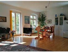 Helles Wohnzimmer mit Essbereich, Holzboden und großen Fenstern mit gelben Vorhängen, die Tageslicht einlassen. Im Raum stehen ein runder Esstisch mit Stühlen, eine Zimmerpflanze, ein niedriger Fernseher, ein Teppich mit Mustern und ein weißer Küchenschrank mit Geschirr.