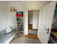 Ein Kinderzimmer mit einem weißen Bücherregal, das Bücher und Spielzeug enthält. Links steht ein Bett mit grauer Sternenbettwäsche und Plüschtieren, im Hintergrund ist eine offene Tür, die in einen Korridor mit Holzboden führt.