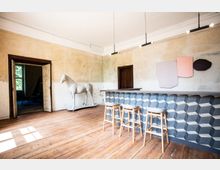 Ein minimalistischer, heller Raum mit Holzboden und gealterten Wänden. Im Vordergrund steht eine moderne Theke mit geometrischem Muster und drei Barhockern, daneben eine weiße Pferdeskulptur; im Hintergrund sind geöffnete Türen und ein Fenster zu sehen.