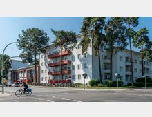 Straßenszene mit einem modernen Wohngebäude aus der Nachkriegszeit, das vier Stockwerke hoch ist und weiße Fassaden mit roten Balkonen hat, umgeben von hohen Bäumen. Im Vordergrund eine Straße mit Radfahrer, Straßenlaternen und parkenden Fahrrädern sowie ein Eingang zur U-Bahn-Station mit blauen Schildern.