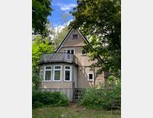 Holzhaus mit Satteldach in einer grünen Umgebung, umgeben von dichten Bäumen. Das Gebäude verfügt über eine verglaste Veranda im unteren Bereich und einen Balkon im oberen Stockwerk.