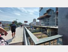 Fußgängerbrücke über Bahngleise in einer städtischen Umgebung, rechts ein modernes Gebäude mit Glasfassade und Gerüstkonstruktionen, links Wohn- und Bürogebäude im Hintergrund. Menschen gehen die Brücke entlang, und unter der Brücke verlaufen mehrere Bahngleise.