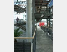 Moderne Innenansicht eines Bürogebäudes mit sichtbarer Stahlkonstruktion und offenen Galerien auf mehreren Ebenen. Rechts verläuft ein langer Gang mit Geländern aus Metall und Holz, links sind Treppen und eine große Palme zu sehen, die einen industriell-minimalistischen Stil ergänzen.