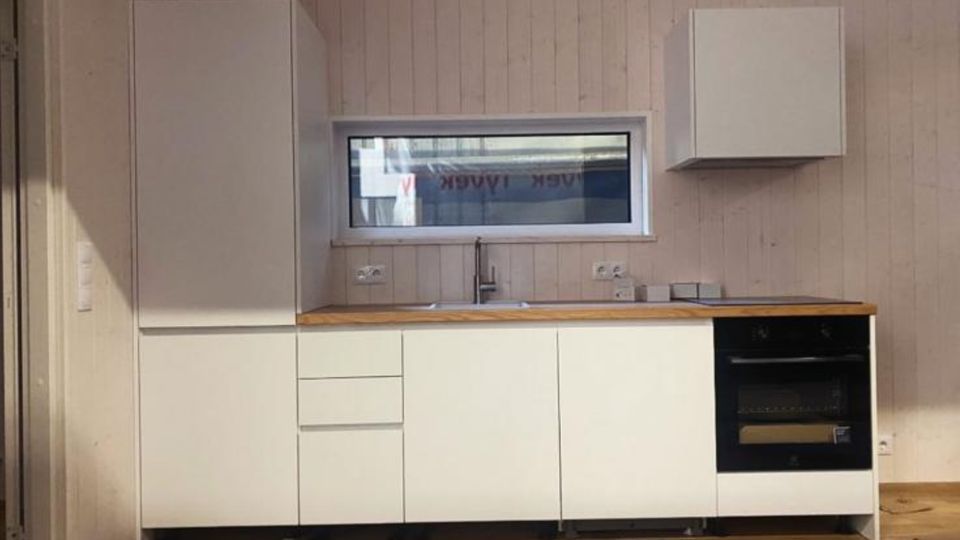 Moderne, minimalistische Küchenzeile mit weißen Schrankfronten und Holzarbeitsplatte vor hell vertäfelter Wand. Unter einem schmalen Fenster befinden sich Spüle und Armatur; rechts ist ein eingebauter Backofen, links ein hoher Hochschrank und darüber ein Hängeschrank.