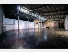 Große, leere Lagerhalle mit industriellem Charakter, gekennzeichnet durch Metallträger, hohe Decken und eine Mischung aus weißen und freiliegenden Ziegelwänden. Der Boden besteht aus glattem Beton, und im Hintergrund sind große Bogenfenster und Backsteinmauern sichtbar.