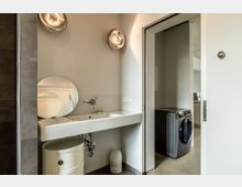 Modernes Badezimmer mit einem weißen Waschtisch, einer runden Spiegelkombination und Wandbeleuchtung. Im angrenzenden Raum ist eine Waschmaschine in einer grauen Nische zu sehen.