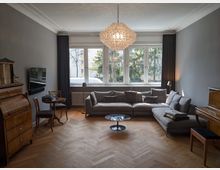 Helles Wohnzimmer mit großem Fensterband und dunklen Vorhängen, Parkett im Fischgrätmuster und einem großen Ecksofa um einen runden Couchtisch. In der Mitte hängt ein großer Kristallleuchter; an den Wänden stehen antike Holzmöbel, ein Fernseher und ein kleiner Tisch mit Stühlen.