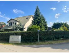 Wohnhaus mit Satteldach und hellbeiger Fassade, umgeben von einer hohen Hecke und Nadelbäumen. Im Vordergrund verläuft eine asphaltierte Straße, daneben befindet sich ein Stromkasten und ein Straßenschild.