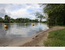 Flussufer mit Sandstrand und angrenzender Grünfläche, gesäumt von Bäumen und Büschen; mehrere gelbe Bojen schwimmen im Wasser, während der Himmel von Wolken und blauem Himmel durchzogen ist.