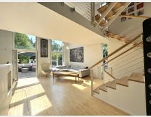 Heller, moderner Wohnbereich mit doppelter Raumhöhe, großen Fensterfronten und offener Treppe mit Holzstufen und Seilgeländer zur Galerie. Links führt eine Glastür auf eine Terrasse, im Raum stehen ein Ecksofa, ein Sessel und ein Couchtisch auf hellem Holzboden.