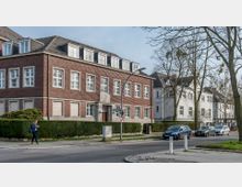 Straßenszene mit einem großen, mehrstöckigen Gebäude aus rotem Klinker und weißen Fenstern, flankiert von weiteren Häusern in hellen Farben. Im Vordergrund eine Straße mit parkenden Autos, einer Fußgängerin und Bäumen am Rand.