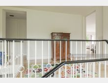 Treppenabsatz eines Wohnhauses mit weißem Geländer, hellem Holzboden, einem bunten Teppich mit Blumenmuster und einem braunen Holzschrank an der Wand; im Hintergrund ein Blick in ein helles Zimmer mit einem Bett.