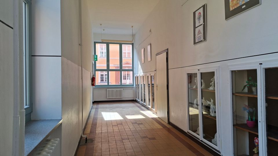 Langer, heller Flur in einem Schul- oder Verwaltungsgebäude mit Fliesenboden, hohen weißen Wänden und großen Fenstern am Ende. Entlang der rechten Seite stehen verglaste Vitrinen mit Ausstellungsobjekten, darüber hängen gerahmte Bilder; am Fenster sind Heizkörper und ein Ausgangsschild zu sehen.