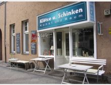 Fassade eines kleinen Cafés mit der Aufschrift „Klötze u. Schinken – Kunst und Raum“ auf einem blauen Schild über der Tür. Vor dem Eingang stehen weiße Holztische und Bänke sowie Tafeln mit Speisenangeboten, die an der beigefarbenen Wand befestigt sind.
