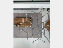 Ansicht eines modernen Wohnzimmers aus der Vogelperspektive. Ein metallischer Kronleuchter im Ast-Design ragt in den Vordergrund, darunter ein Holztisch mit Büchern und Fernbedienung auf einem beigefarbenen Teppich; weiße Dielenboden und ein graues Sofa sind im Hintergrund sichtbar.