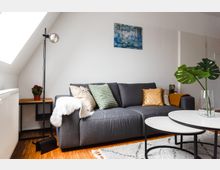 Wohnzimmer mit einer grauen Sofa-Ecke, dekoriert mit bunten Kissen und einer weißen Decke. Daneben steht ein Beistelltisch mit einer Zimmerpflanze, während im Vordergrund zwei runde, ineinander verschachtelte Couchtische und eine Vase mit grünen Blättern zu sehen sind. Ein Bild mit Naturmotiv hängt an der weißen Wand.