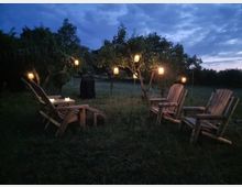 Ein Garten bei Dämmerung mit vier Holzstühlen und einem kleinen Holztisch auf einer Wiese. Rundherum hängen mehrere leuchtende Laternen an den Ästen der Obstbäume.