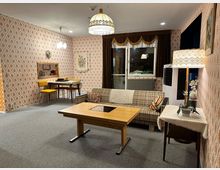 Wohnzimmer im Retro-Stil mit gemusterten Tapeten in Orange- und Beigetönen. Der Raum enthält eine Polstercouch mit kariertem Muster, einen Holztisch, eine Stehlampe mit floralem Lampenschirm, sowie einen kleinen Esstisch mit gelben Stühlen und einer Durchreiche in der Wand.