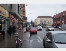 Straßenszene in einer belebten Stadt mit mehrstöckigen Gebäuden auf beiden Seiten, darunter Geschäfte, Restaurants und Supermärkte. Im Hintergrund ist der Eingang zur U-Bahn-Station "Hermannstraße" zu sehen, mit geparkten Autos, Fahrradständern und Fußgängern entlang des Gehwegs.