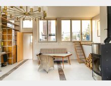 Wohnzimmer mit minimalistischem Design, großen Fenstern und einer kleinen Holzleiter, die zu einem erhöhten Bereich führt. Im Raum befinden sich eine graue Couch, ein Holztisch, ein Korbstuhl, ein Kaminofen mit gestapeltem Holz daneben und ein Metallregal mit Holzverkleidung.