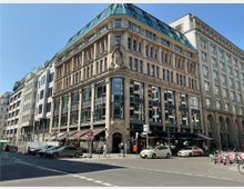 Straßenecke mit einem historischen Gebäude im neoklassizistischen Stil, gekennzeichnet durch Säulenfassaden und große Fenster. Im Erdgeschoss befinden sich Geschäfte und Restaurants, davor parkende Autos und Fahrräder, umgeben von weiteren Büro- und Wohngebäuden.