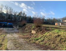 Offene Grünfläche mit vereinzelten Büschen und grasbewachsenen Hügeln, im Hintergrund mehrstöckige Wohngebäude mit blauen Dächern sowie ein kleines, eingeschossiges Gebäude aus Ziegelstein. Links vorne ist ein gepflasterter Weg und einige geparkte Baugeräte sichtbar.