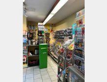 Kleiner Zeitschriftenladen mit engen Gängen, gefüllt mit Regalen voller Magazine, Zeitungen und Tabakwaren. Im Hintergrund eine grüne PIN-Postbox sowie dekorative Poster an der gelb gestrichenen Wand.