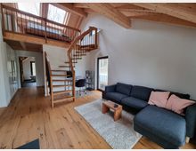 Offenes Wohnzimmer mit Holzdecke und sichtbaren Dachbalken, einer Wendeltreppe aus Holz und Metall, einer grauen Eckcouch mit rosa Kissen, einem kleinen Holztisch und einem Arbeitsbereich mit Schreibtisch und Stuhl. Große Fenster und Oberlichter lassen viel Tageslicht herein, der Boden besteht aus Holz.