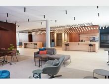 Ein moderner Innenbereich eines Büros oder Gemeinschaftsraums mit offenen Sitzbereichen, hellem Holzdekor und einer großen Theke im Hintergrund. Die Decke ist mit einer Vielzahl von kleinen Lichtern dekoriert, und der Raum enthält Sitzmöbel wie Sofas, Stühle und Tische in neutralen Farbtönen.