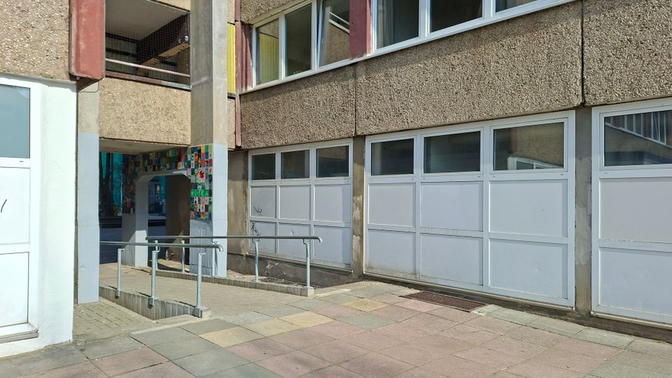 Außenansicht eines mehrstöckigen Betongebäudes mit rauer Fassade und großen, weiß gerahmten Fenster- und Türpaneelen im Erdgeschoss. Vor dem Eingang führt eine Rampe mit Metallgeländer über einen gepflasterten Gehweg; neben dem Durchgang ist eine bunte, mosaikartige Wandfläche zu sehen.