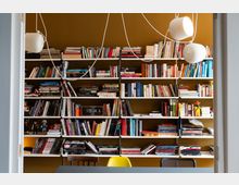 Ein Raum mit Bücherregalen, die bis zur Decke reichen und mit einer Vielzahl von Büchern, Magazinen und kleinen Figuren gefüllt sind, vor einer senfgelben Wand. Im Vordergrund steht ein Tisch mit einer gelben und einer hölzernen Stuhllehne sichtbar, darüber hängen minimalistische, weiße Hängeleuchten.