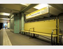 U-Bahnhof mit gelber Sitzbank, beschriftet mit "Viktoria-Luise-Platz" auf einem Schild der Linie U4. Der Bereich zeigt eine metallene Stütze, einen türkisfarbenen Türrahmen und helle Kacheln, mit einem beleuchteten Ausgang Richtung Treppe im Hintergrund.