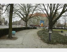 Stadtpark mit gepflasterten Wegen, großen Bäumen und einer Rasenfläche, die von Sträuchern umgeben ist; im Hintergrund mehrgeschossige Gebäude mit Geschäften und Werbeplakaten.