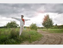 Eine Vogelscheuche steht auf einer grünen Wiese mit wilden Blumen, neben einem unbefestigten Weg, der durch eine ländliche Landschaft mit Bäumen und Feldern führt. Der Himmel ist bewölkt, mit leichtem Sonnenlicht im Hintergrund.